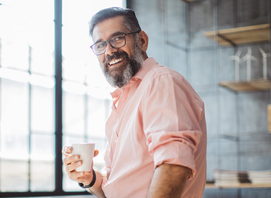 Man with glasses smiling at camera with coffee in hand.